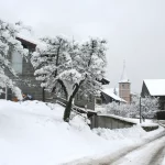 Rue de Cervens bordée de maisons et d’arbres enneigés avec le clocher en arrière-plan
